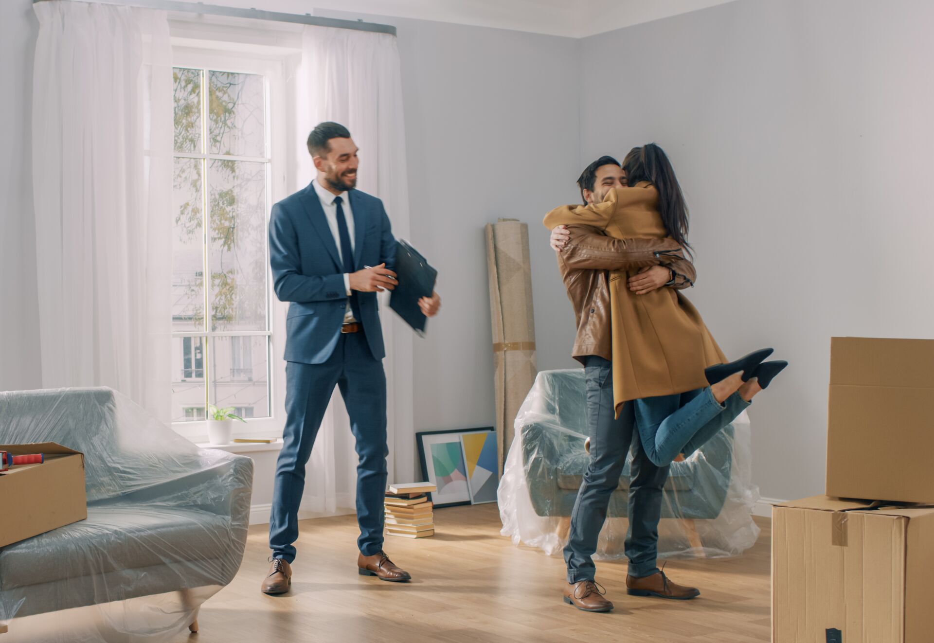 Freudige Hausübergabe durch SCHULER INVESTMENTS: Junges Paar umarmt sich in neuem Zuhause, Makler im Hintergrund. Umzugskartons sichtbar.