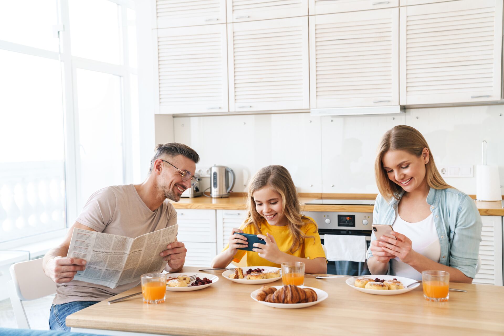 Familie frühstückt entspannt: Vater liest Zeitung, Tochter spielt am Handy, Mutter tippt. SCHULER INVESTMENTS im Hintergrund denkbar.