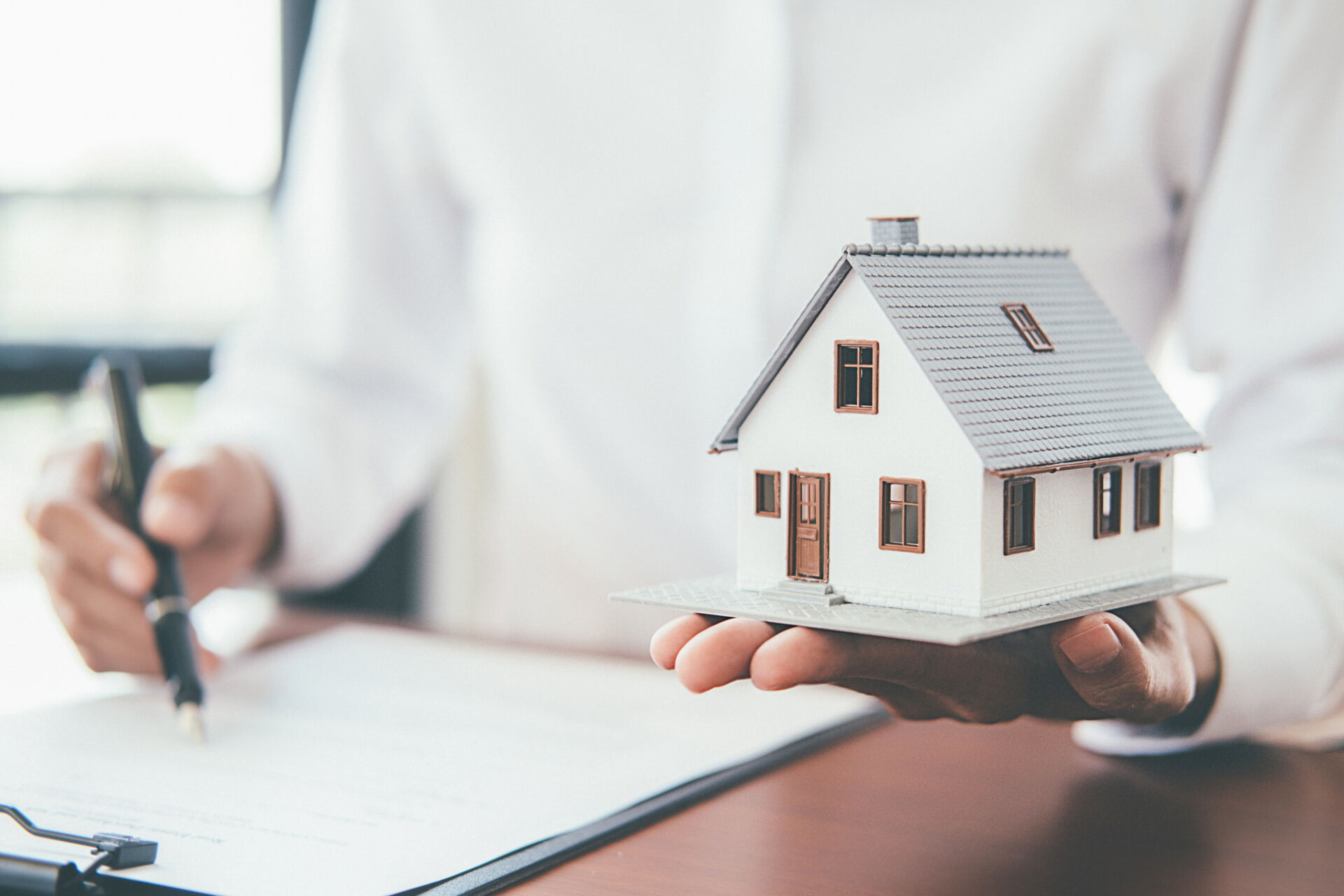 Person hält Miniaturhaus, unterschreibt Dokument. Fokus auf Hausversicherung durch SCHULER INVESTMENTS. Schutz für Ihr Zuhause.