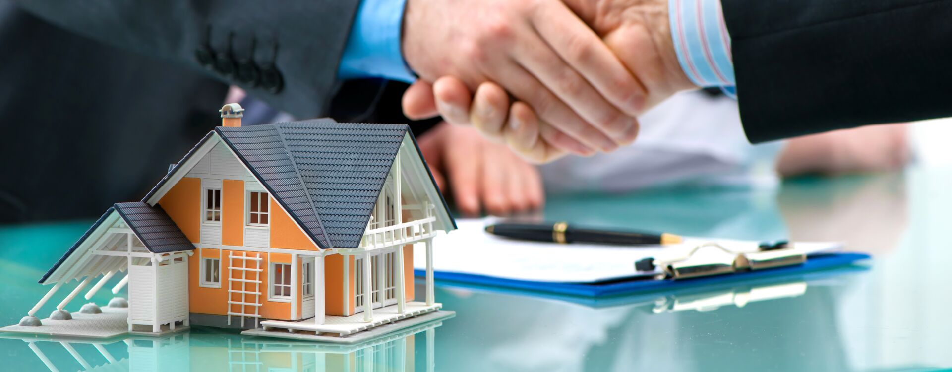 Handschlag beim Hausverkauf, Schlüsselübergabe im Hintergrund. Miniaturhaus im Vordergrund. SCHULER INVESTMENTS als Ihr Partner.