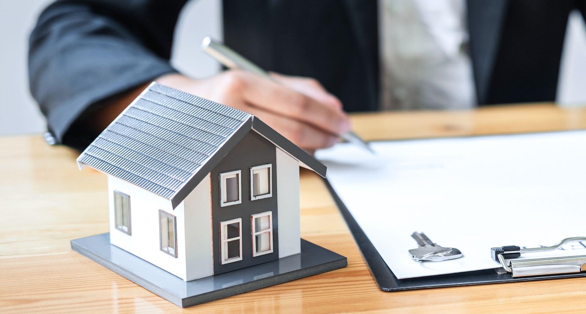 Person rechnet und unterschreibt Vertrag neben Hausmodell und Schlüssel. Fokus auf Immobilienfinanzierung durch SCHULER INVESTMENTS.