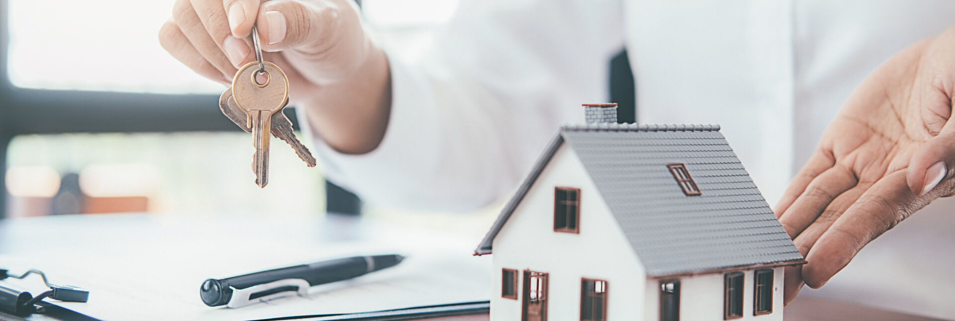 Hände präsentieren Schlüssel neben einem Hausmodell und einem Vertrag. Symbol für Immobilienübergabe durch SCHULER INVESTMENTS.